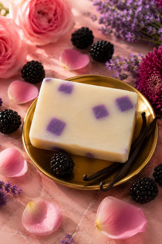Bar of soap with purple specks on a gold plate surrounded by flowers, blackberries, and vanilla beans.