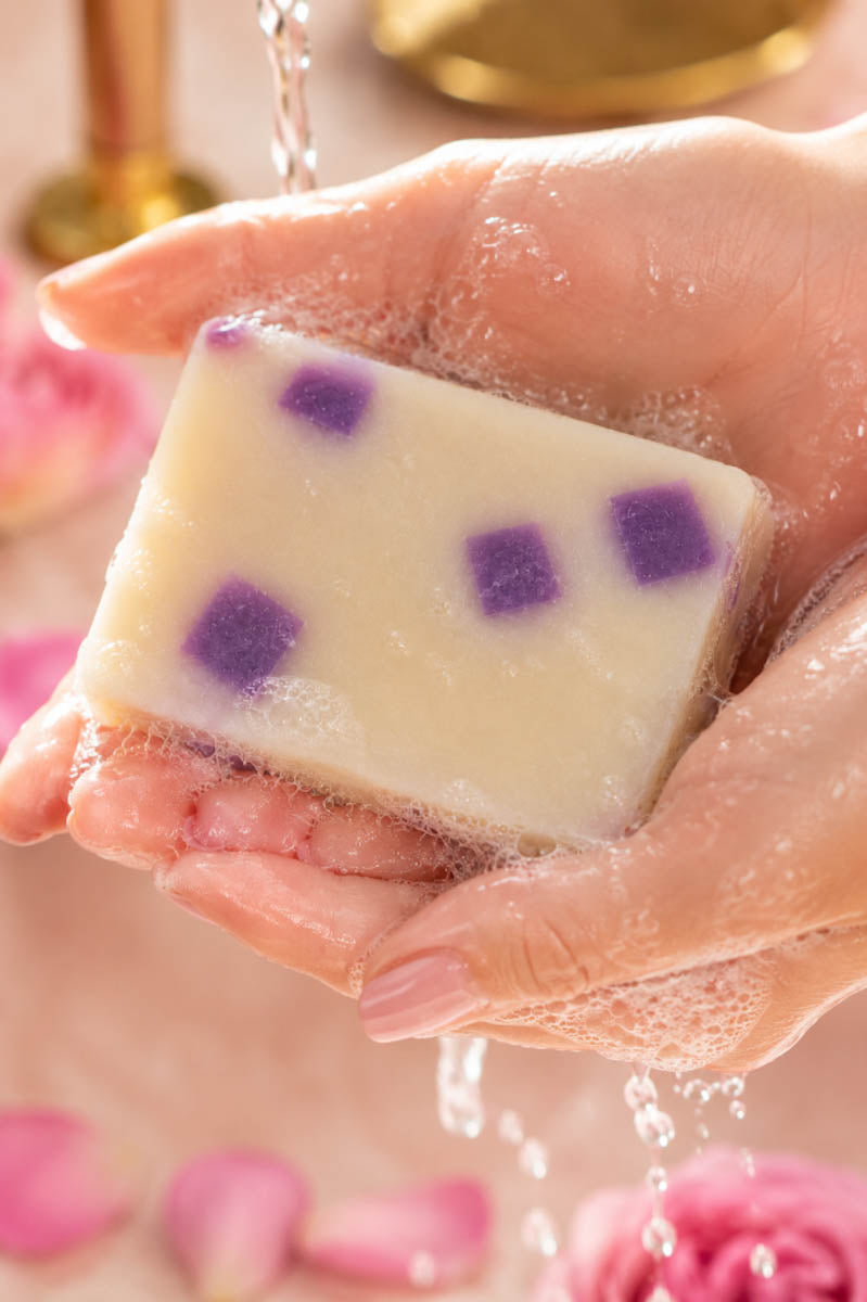 Hand holding a bar of Honeysuckle Lifestyle soap with purple spots in a bathroom setting