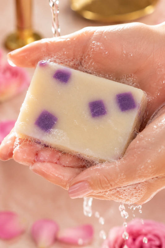 Hand holding a bar of Honeysuckle Lifestyle soap with purple spots in a bathroom setting